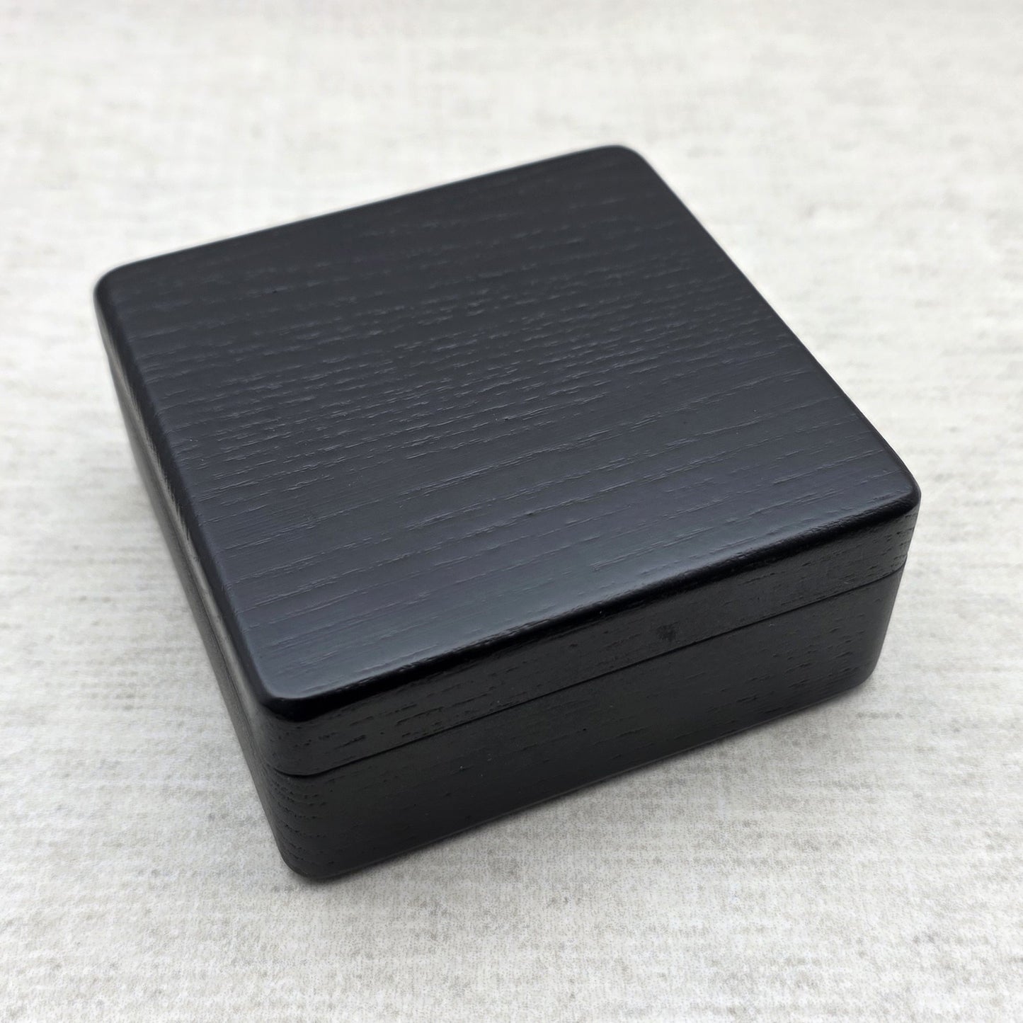 Closed black painted ash wood gift box showing sleek rounded corners and natural wood grain texture under light, modern minimalist design.