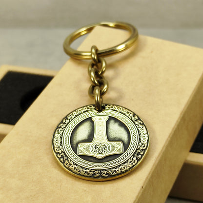 Close-up of a brass Thor's Hammer coin keychain with deep laser engraving, antique patina finish, and decorative border on a craft gift box.