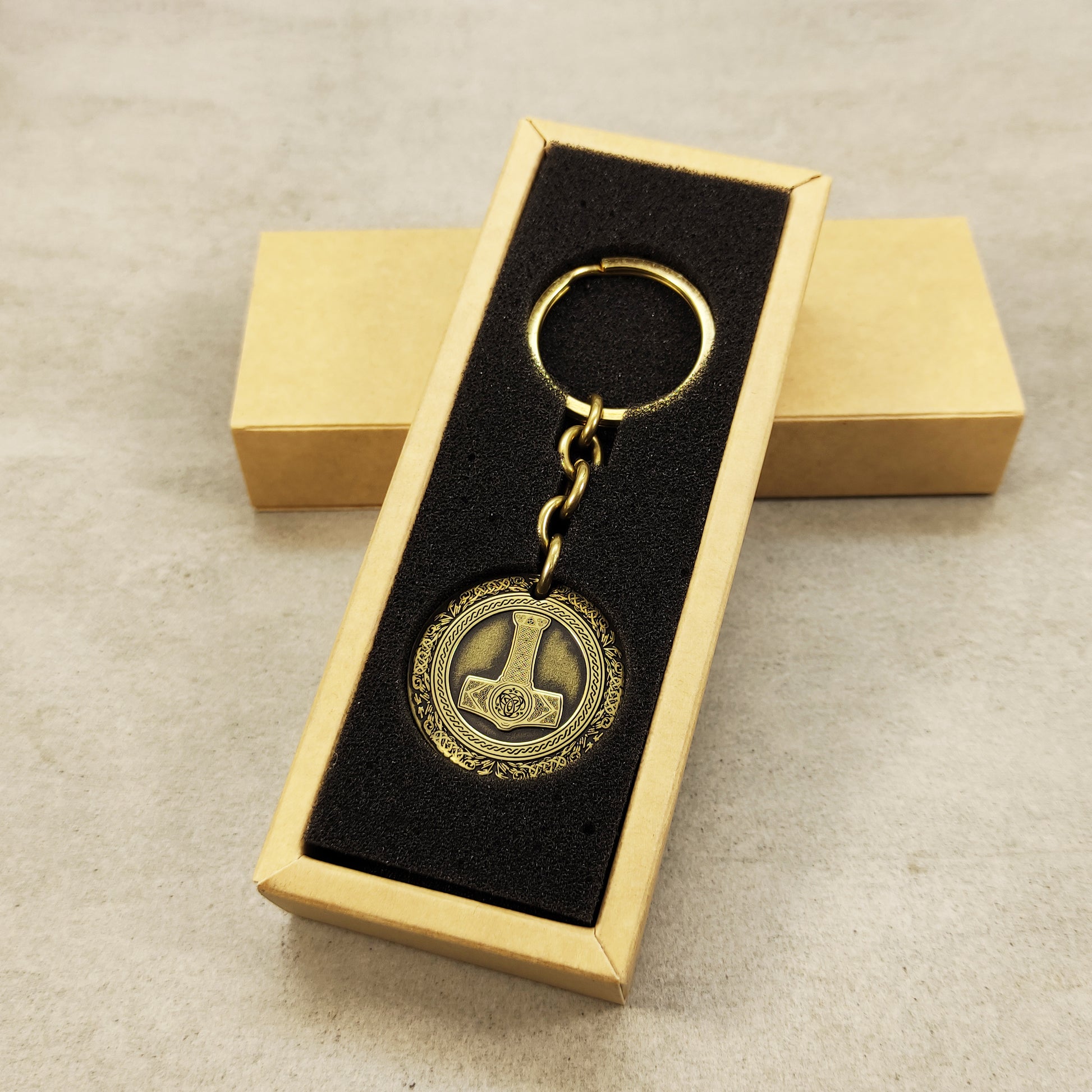 Vertical view of a brass Mjolnir coin keychain in a premium black gift box insert, showcasing the polished brass surface and engraved viking details.
