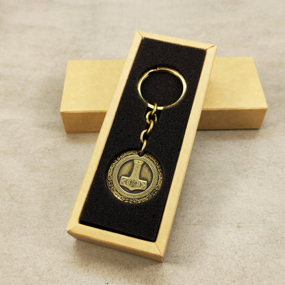 Vertical view of a brass Mjolnir coin keychain in a premium black gift box insert, showcasing the polished brass surface and engraved viking details.