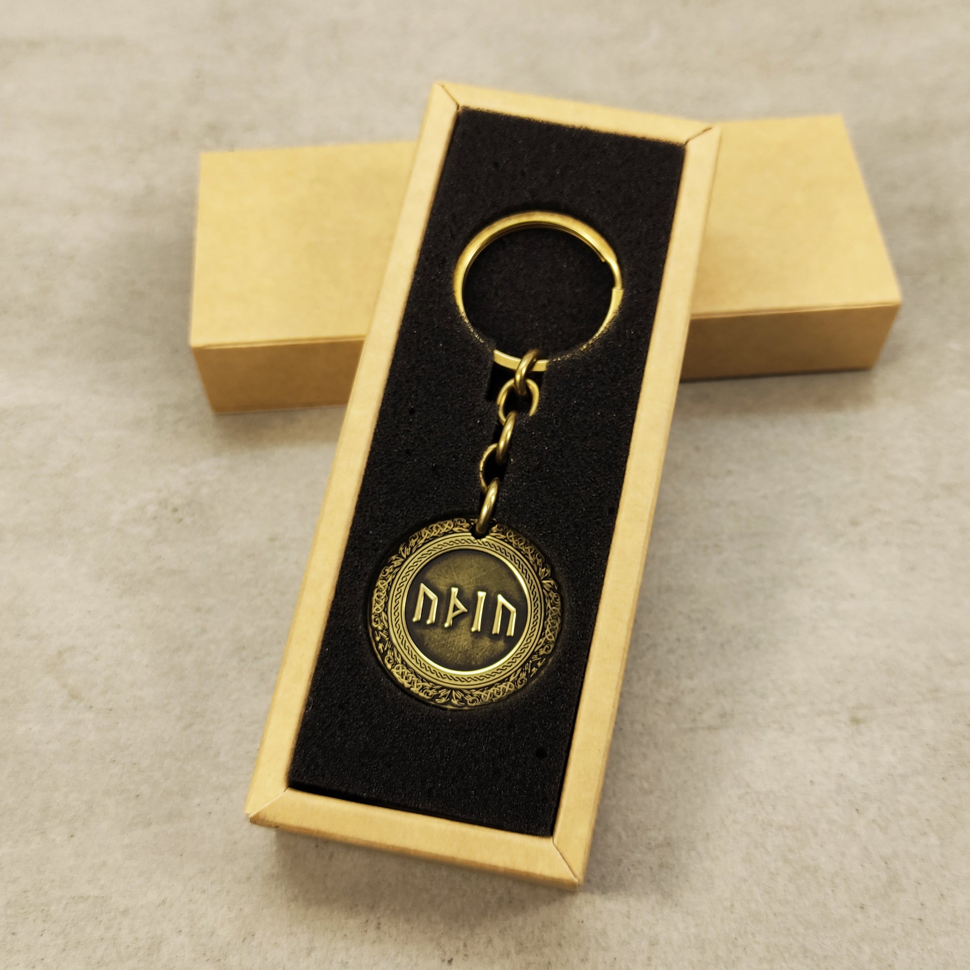 Vertical view of a brass rune coin keychain in a premium black gift box insert, showcasing the gold-tone metal and dark engraved details.
