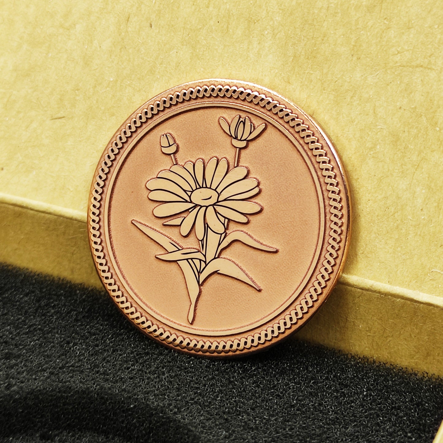 Personalized copper Daisy coin leaning against a square gift box, showing the warm copper tone and precision of the floral laser etched design.